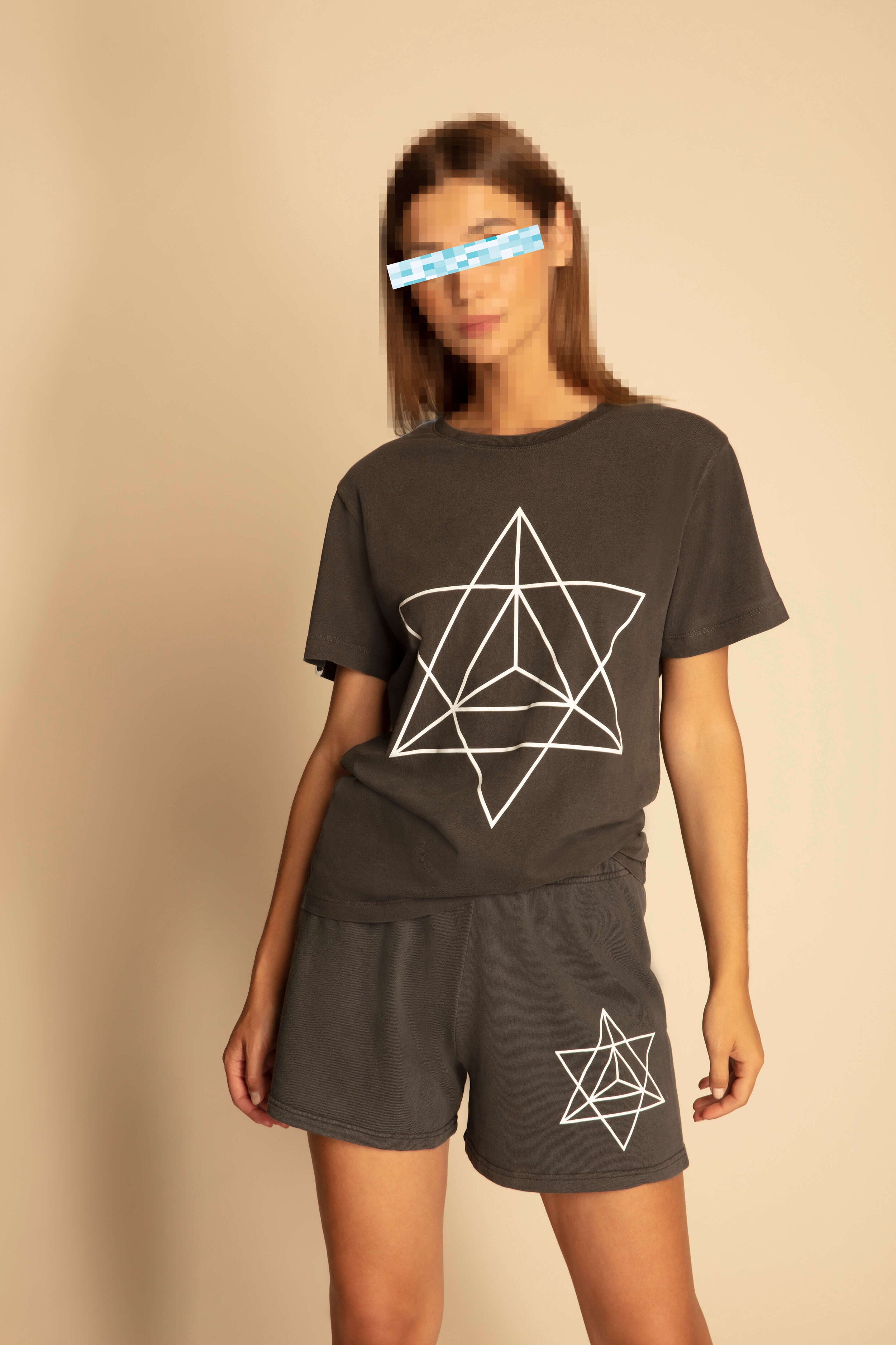A person stands against a plain background, wearing a dark GFLApparel T-shirt and Merkaba Shorts in Space Glow with geometric star designs inspired by Sacred Geometry. Crafted from breathable cotton, their attire blends style and comfort seamlessly. For privacy, their face is pixelated.