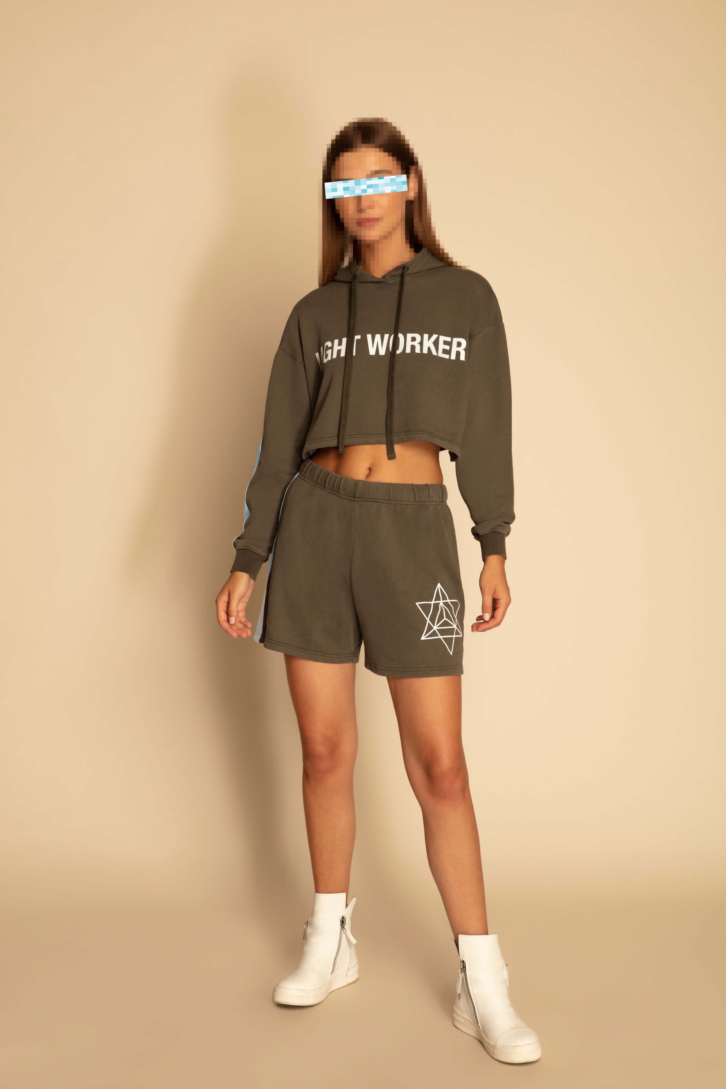 Person standing against a plain background wearing a GFLApparel Light Worker Cropped Hoodie in Caladan, matching Merkabah shorts with a star design, and white ankle boots.