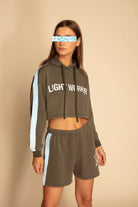 A person stands against a beige background, wearing a Light Worker Cropped Hoodie in Caladan by GFLApparel and matching Merkabah shorts, both with white side stripes. Their face is digitally blurred to protect identity.