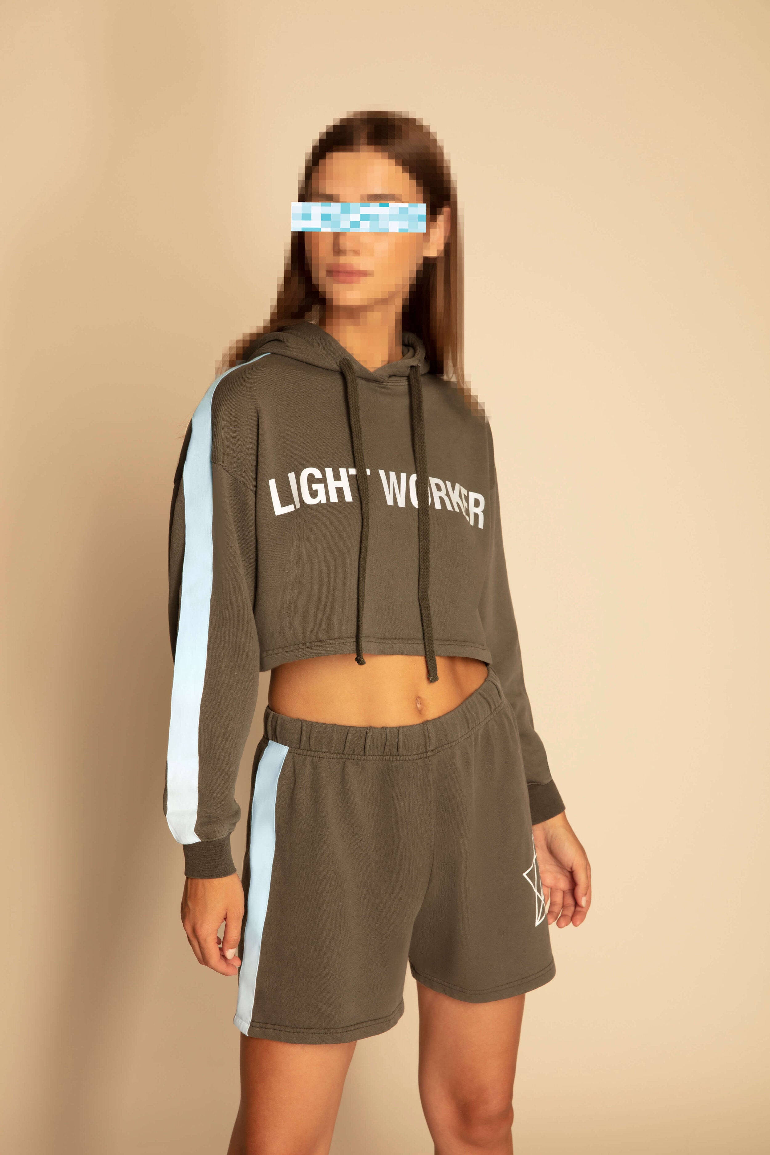A person stands against a beige background, wearing a Light Worker Cropped Hoodie in Caladan by GFLApparel and matching Merkabah shorts, both with white side stripes. Their face is digitally blurred to protect identity.