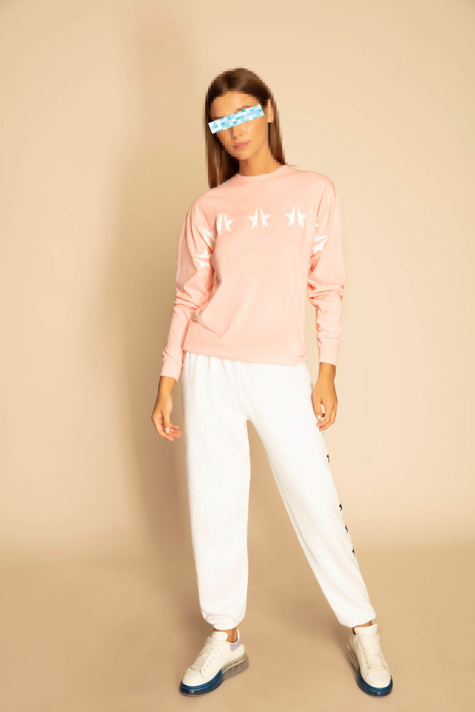 A person stands against a beige background wearing a pink GFLApparel GFL Stars L/S in Sun Fade with star designs, white pants, and white sneakers. Their eyes are covered by a blue, pixelated effect. The relaxed fit of the breathable cotton shirt adds a comfortable vibe to the look.