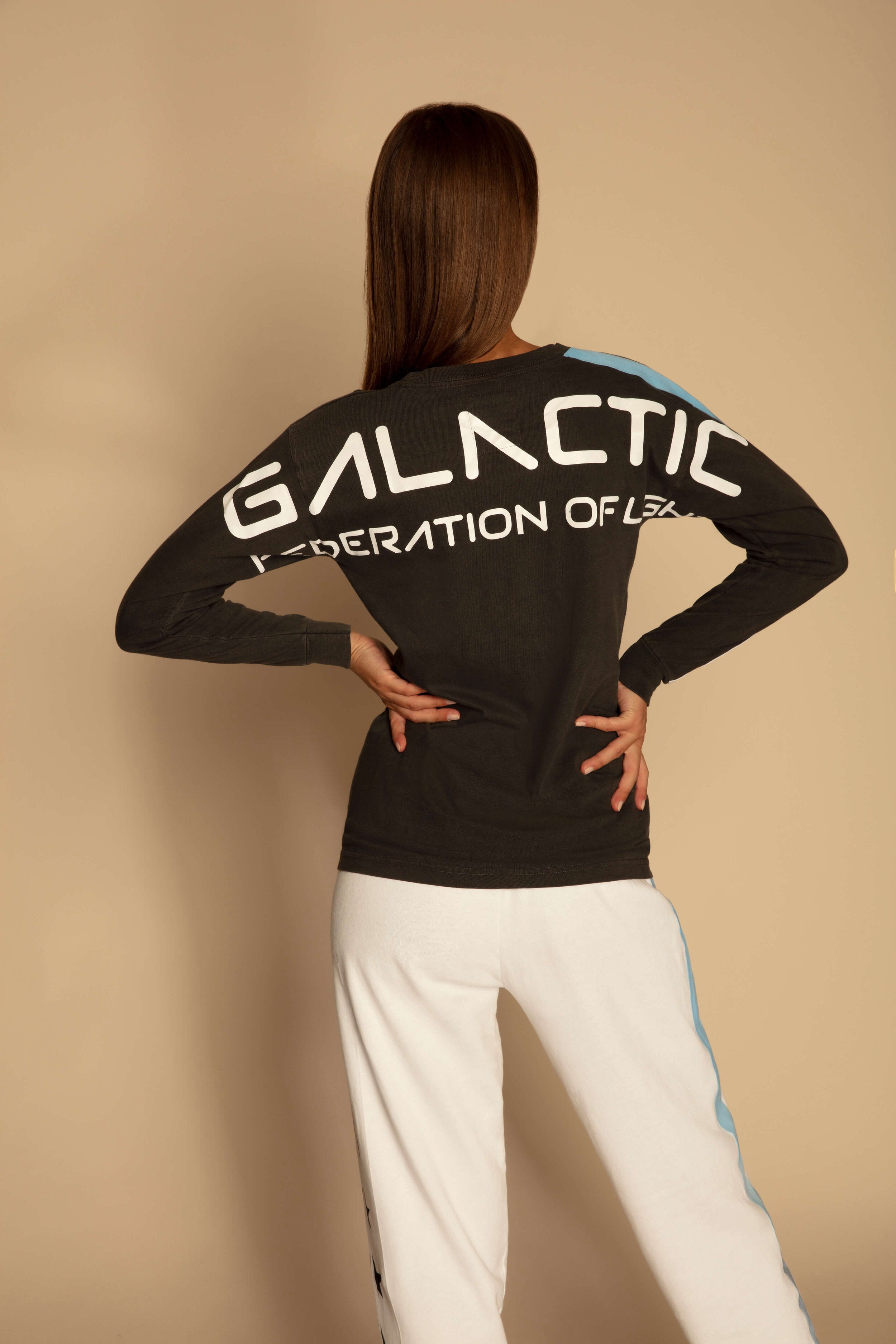 Person with long hair wearing a breathable cotton black GFLApparel GFL Stars L/S in Space Glow shirt that says "GALACTIC Federation of L3X" on the back, and white pants with a blue stripe, stands with hands on hips facing away.